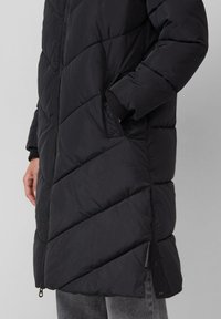 Zwarte pufferjas met een gequilt ontwerp en chevronpatronen, zijzakken en ribgebreide manchetten. Ritsluiting loopt door tot de zoom.