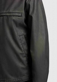 Čierna kožená bunda s náprsným vreckom s lemom, bočným vreckom a dlhým rukávom na svetlosivom textúrovanom pozadí.