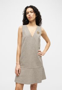 Γυναίκα με σγουρά μαλλιά, φοράει αμάνικο μπεζ και μαύρο φόρεμα με καρό σχέδιο, λαιμόκοψη σε σχήμα V και μια χρυσή καρφίτσα σε σχήμα λουλουδιού.