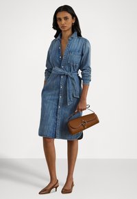 Moteris dėvi džinsinę suknelę su diržu, rankoje laiko rudos odos rankinę ir dėvi rudas žemas aukštakulnes su užkulniu sulenktas basutes, stovėdama ant neutralios spalvos fono.