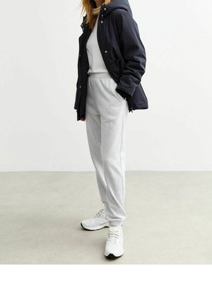 Personne portant une tenue de sport décontractée avec une veste à capuche noire, un sweat-shirt blanc, un pantalon de jogging gris et des baskets blanches, debout sur un sol gris.