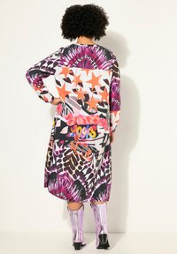 Langes Kleid mit lebhaften mehrfarbigen Mustern, darunter Sterne und Tierdrucke. Kombiniert mit kniehohen glänzenden rosa Stiefeln.