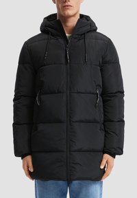 Mann trägt eine schwarze gesteppten Pufferjacke mit Kapuze, vorderem Reißverschluss und seitlichen Reißverschlusstaschen, kombiniert mit blauen Jeans.