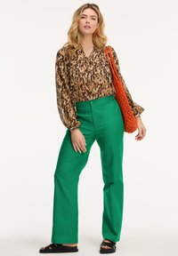 Blusa a maniche lunghe con motivo in marrone e beige, abbinata a pantaloni verdi a vita alta. Presenta una borsa intrecciata arancione e sandali neri.