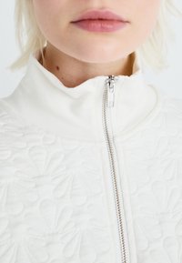 Nærbillede af en person iført en hvid tekstureret jakke med en sølvfarvet lynlås, der er lynet helt op til halsen.