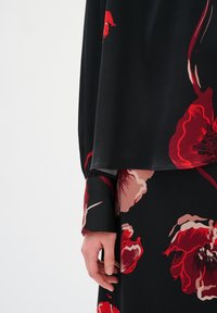 Mano di donna accanto a un tessuto nero con grande stampa floreale rossa e rosa, che mostra i dettagli del polsino della manica e dell'orlo della gonna.