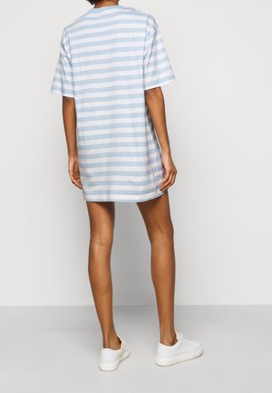 Personne portant une robe à manches courtes, longueur genou, avec des rayures horizontales bleu clair et blanc, associée à des baskets blanches, debout contre un mur blanc.