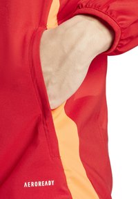 Rote Jacke mit einem orangefarbenen Akzent, ausgestattet mit einer Seitentasche und elastischen Bündchen. Das Material wirkt glatt und leicht, mit Branding am Saum.