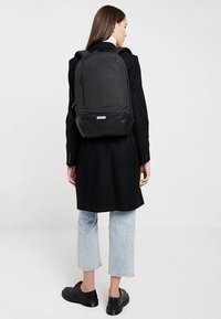 Zaino nero con una finitura opaca e liscia. Presenta una tasca con zip e una base arrotondata. Indossato sopra un cappotto scuro e jeans chiari.