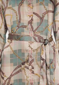 Skjorta med rutmönster som har kedjor och läderremmar. Tillverkad av ett slätt tyg, med knappar, midja med bälte och jordnära, dämpade färger.