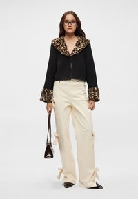 Casaco preto cropped com colarinho de pele sintética de padrão de leopardo, combinado com calças creme com detalhes de laço. Sapatos pretos brilhantes de bico afilado.