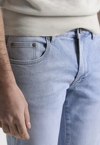 Jeans de mezclilla azul claro con diseño estándar de cinco bolsillos, cerraduras de botón metálico y una textura suave. Mano descansando sobre la cintura.
