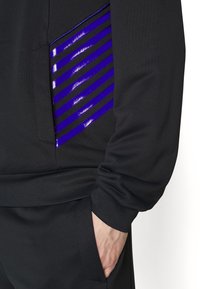 Svart sweatshirt med en sidficka som har glänsande lila ränder. Mjukt tyg, avslappnad passform, elastiska ärmslut och midjeband för komfort.