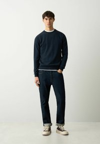 Marineblaues Sweatshirt mit Rundhalsausschnitt und gerippten Bündchen; über einem weißen Hemd getragen. Kombiniert mit dunkelblauen Jeans und braunen Sneakers mit Cremesohlen.