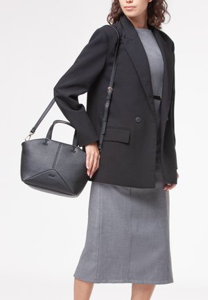 Borsa a mano in pelle nera con finitura testurizzata, dotata di manici superiori doppi e una tracolla removibile, portata da una persona in blazer e gonna.