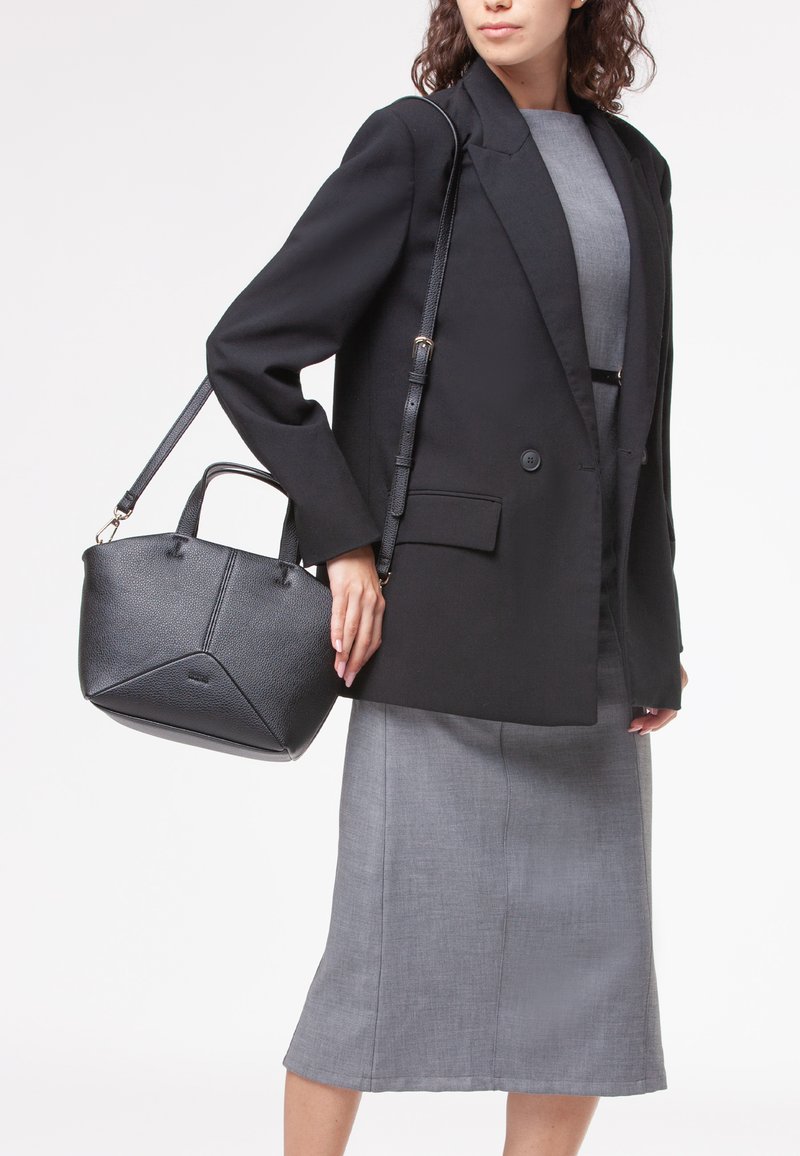 Borsa a mano in pelle nera con finitura testurizzata, dotata di manici superiori doppi e una tracolla removibile, portata da una persona in blazer e gonna.