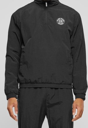 Man wearing a black long-sleeve sports jacket with a front half-zip and a circular "Karl Kani Sport" logo on the chest.