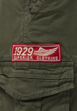 Tissu vert avec un patch rectangulaire rouge affichant en blanc le texte "1929 Superior Clothing" et un motif d'aile au-dessus d'une poche pliée.