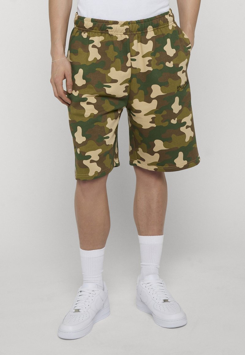 Homme portant un short camouflage vert, marron et beige, des chaussettes blanches montantes et des baskets blanches, debout les mains dans les poches devant un fond gris.