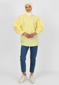 Tunique en coton jaune avec des manches bouffantes et un col haut, associée à un jean en denim bleu et des baskets beige clair. Coupe simple et décontractée.