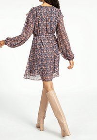 Robe semi-transparente à motif floral en bleu marine et pêche, avec des manches à volants et une taille cintrée. Associée à des bottes nude montant jusqu'au genou.