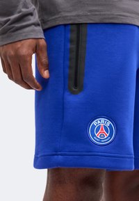 Άνδρας φοράει μπλε σορτς Paris Saint-Germain με μαύρη λεπτομέρεια φερμουάρ και γκρι μπλούζα με μακριά μανίκια, χέρι χαλαρό στο πλάι.