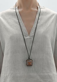 Blouse rayée gris clair avec un décolleté en V. Un collier en corde noire présente un pendentif carré aux tons terreux et à motif géométrique.
