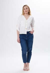Weiße Kurzarmbluse mit Knopfverschluss und zwei Brusttaschen, kombiniert mit einer dunkelblauen Jeans und weißen Riemchensandalen. Glatte Stofftextur.