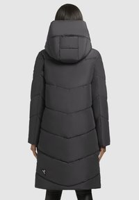 Mustas puffer-jakk, millel on chevron-kvilditud disain, suur kapuuts ja lukuga taskud, millel on logoplekk all vasakul küljel.