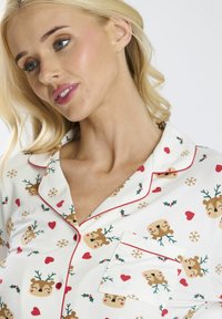 Pigiama in cotone bianco con una stampa festiva di renne e cuori. I bordi rossi accentuano il colletto e la tasca. Tessuto morbido e liscio.