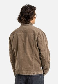 Chaqueta de pana marrón con un corte relajado, que presenta un cuello subido, puños abotonados y dos botones en la parte posterior. Tejido texturizado, sin patrones visibles.