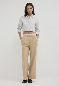 Camicia a righe accorciata con maniche lunghe, pantaloni beige a gamba larga e scarpe nere. I materiali sembrano leggeri; le righe sono verticali.