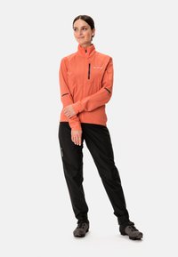 Lätt orange jacka med hög krage och dragkedjeficka, kombinerad med svarta byxor och grå skor, bärs av en person som står med armarna korsade.