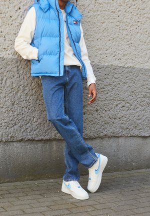 Persoon die een blauwe donsbodywarmer, een crèmekleurige trui, een blauwe spijkerbroek en witte sneakers met lichtblauwe swooshes draagt, staand tegen een getextureerde muur.