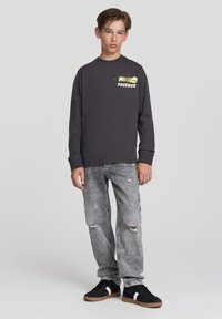 Szara koszula z długim rękawem z grafiką Pokémona na piersi, w połączeniu z podartymi szarymi dżinsami i czarnymi trampkami z białymi paskami.