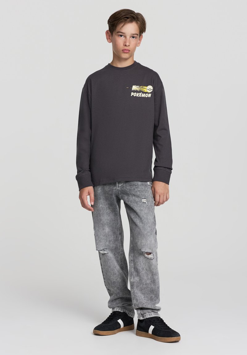Szara koszula z długim rękawem z grafiką Pokémona na piersi, w połączeniu z podartymi szarymi dżinsami i czarnymi trampkami z białymi paskami.
