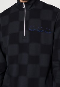Pullover nero con zip a quarto realizzato in tessuto leggero con motivo a quadri. Presenta una zip silver e testo ricamato blu sul petto.