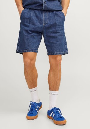 Bermuda in denim con vestibilità rilassata, di colore blu, con tasche frontali, orli risvoltati e cuciture visibili, abbinati a sneaker blu e calzini bianchi.