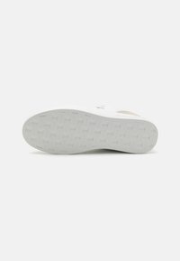 White sneaker with a textured rubber sole, featuring a beige suede accent on the upper and embossed branding for added detail.