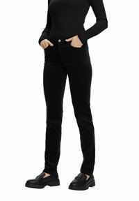 Personne portant un pantalon slim noir, un haut à manches longues côtelé noir et des mocassins noirs, debout les mains dans les poches.