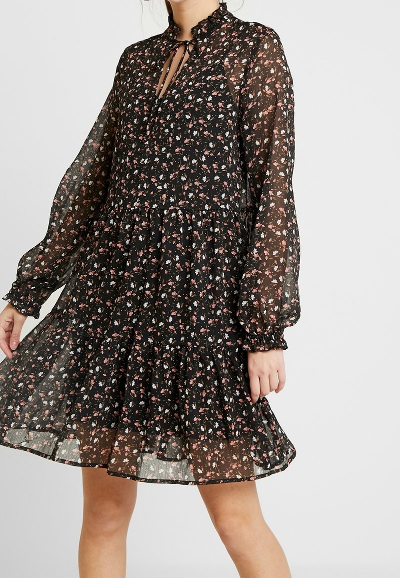 Femme portant une robe noire transparente à manches longues avec imprimé floral rose et blanc, col à volants, jupe fluide et poignets froncés.