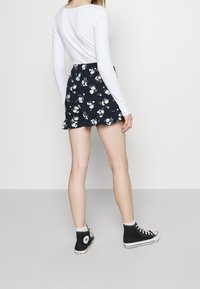 Dunkelblaue Shorts mit floralem Muster und einem Rüschensaum, kombiniert mit einem langärmligen weißen Oberteil und schwarzen High-Top-Sneakern.