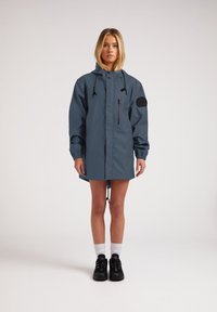 Chaqueta impermeable azul con capucha, mangas largas y dobladillo ajustable. Cuenta con un bolsillo con cremallera y un parche negro del logo en la manga.