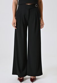 Zwarte wijde broek met een platte tailleband, zijplooien en een decoratieve ronde gesp. Glad oppervlak en lichtgewicht stof.