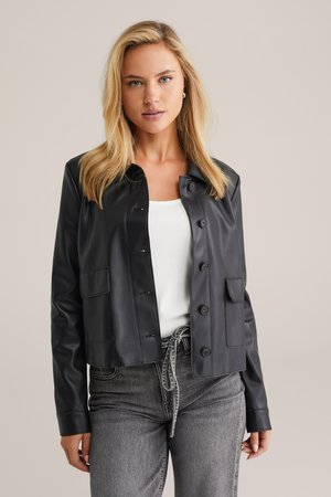 Zwarte nepleer jas met drukknoopsluitingen, twee voorzakken en een cropped ontwerp. Gedragen over een witte top en grijze jeans.