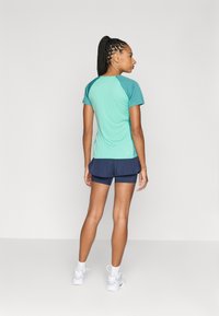 Žena s pleteným ohonem, oblečená v teal krátkém rukávu, námořnických sportovních šortkách, bílých ponožkách a bílých teniskách, stojící proti jednolitému pozadí.