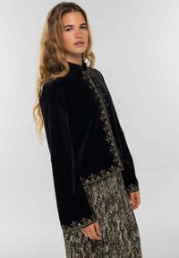 Veste en velours noir avec broderie dorée le long des bords, dotée de manches longues et d'un col montant, portée sur une jupe à motifs.