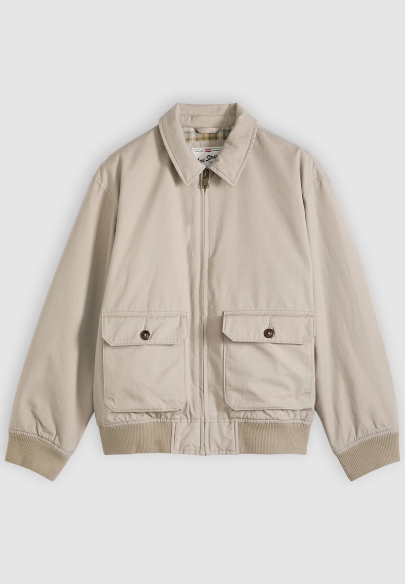 Giacca beige con zip, colletto appuntito, due tasche anteriori con pattina e bottoni, polsini e orlo a coste, distesa su uno sfondo bianco.
