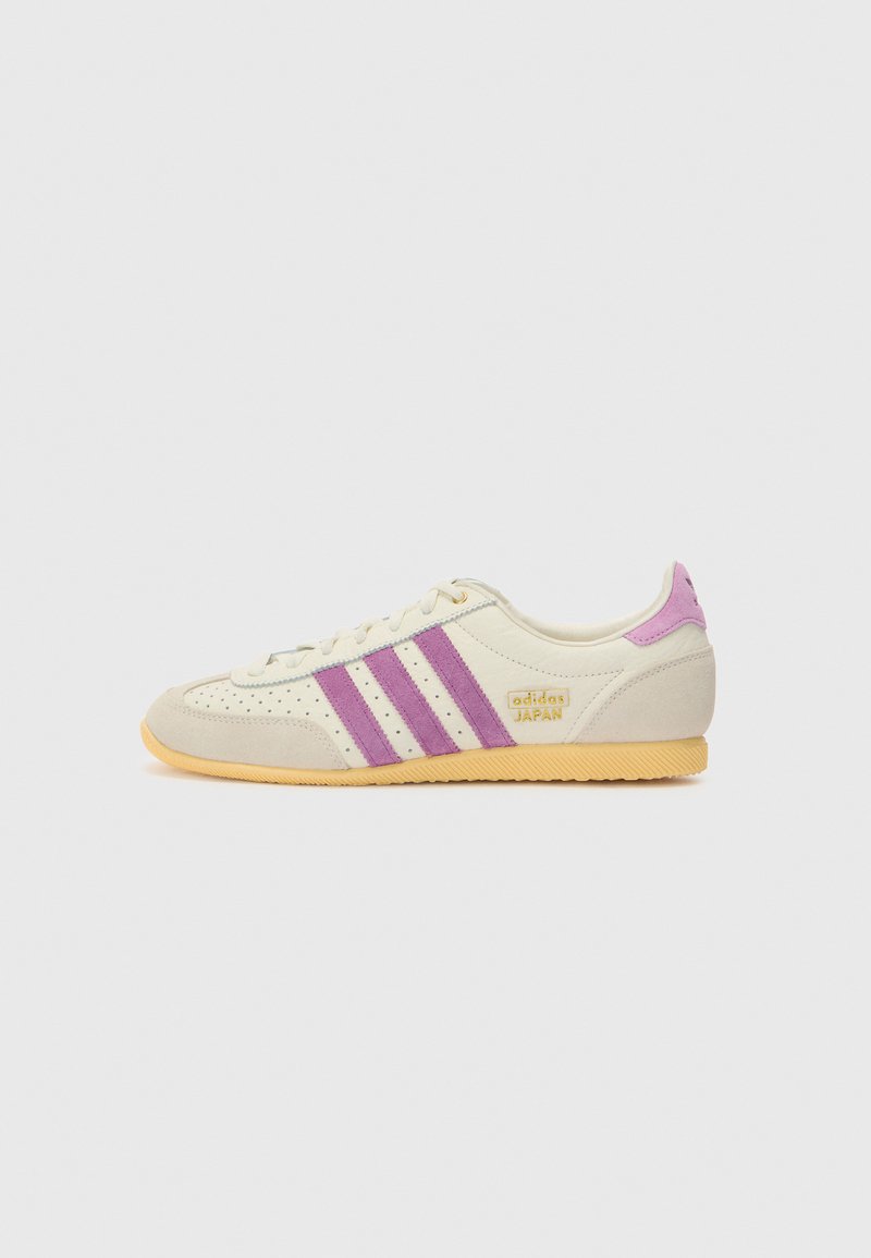 Vit sportskor med lila ränder, mockadetaljer och perforerade ytor. Har en beige gummisul och en guldsymbol med texten "adidas JAPAN."