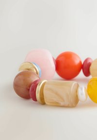 Krāsainā pērļu aproce, kas ietver apaļas un ģeometriskas formas, izgatavota no materiāliem, piemēram, stikla un zeltainām akcentiem, rozā, oranžā un dzeltenā toņos.